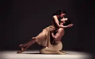 Två balettdansare i en dynamisk pose på en scen med mörk bakgrund, vilket framhäver deras rörelse och uttryck.