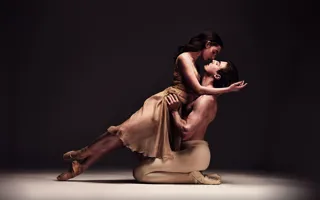 Två balettdansare i en dynamisk pose på en scen med mörk bakgrund, vilket framhäver deras rörelse och uttryck.