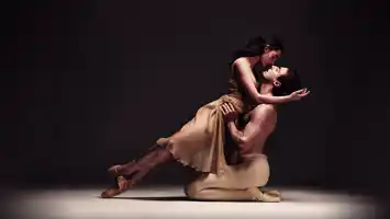 Två balettdansare i en dynamisk pose på en scen med mörk bakgrund, vilket framhäver deras rörelse och uttryck.