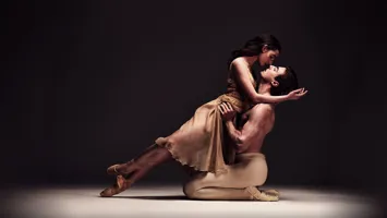 Två balettdansare i en dynamisk pose på en scen med mörk bakgrund, vilket framhäver deras rörelse och uttryck.