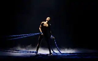 En man står mitt på en mörk scen, han är bunden både runt benen och armarna med tjocka rep.