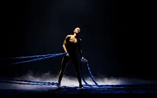 En man står mitt på en mörk scen, han är bunden både runt benen och armarna med tjocka rep.