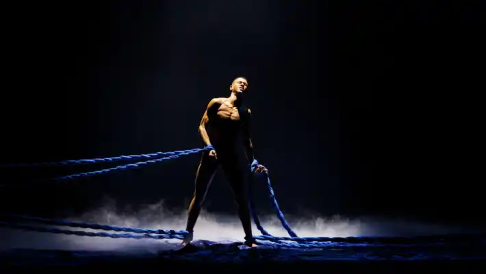 En man står mitt på en mörk scen, han är bunden både runt benen och armarna med tjocka rep.