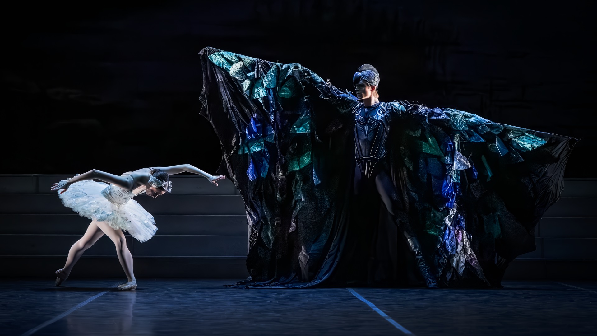 A man dressed in a bird-like cloak spreads his arms in front of a ballerina who bends down.