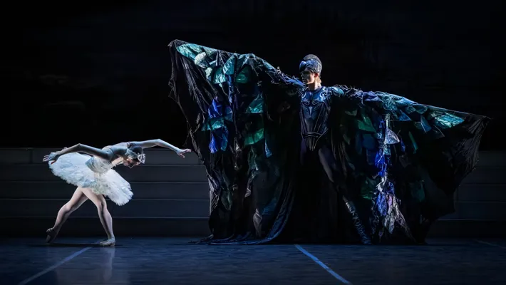 A man dressed in a bird-like cloak spreads his arms in front of a ballerina who bends down.