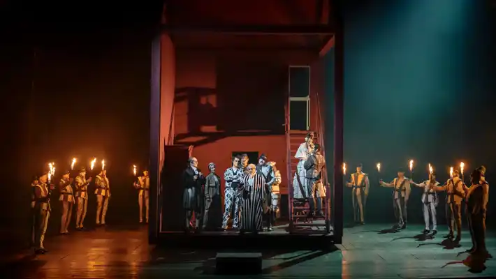 In a large stage room, men with torches stand in a semicircle around a red house with several people inside.