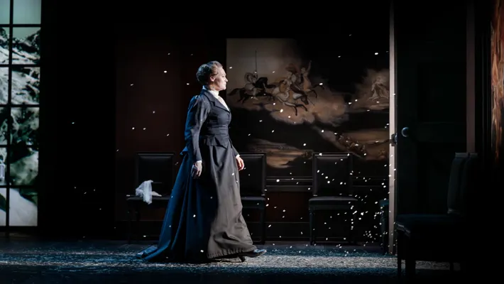 A woman in a dark, long dress stands in the light of a doorway, snow swirling around her.