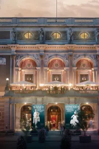 A large facade with trees in front bathed in summer evening light, a church in the background.