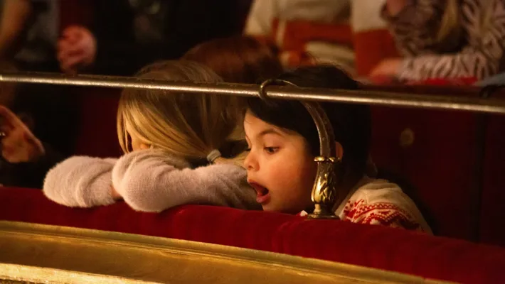 Person with blond hair rests his head on the ledge in a theater - a second person looks open-mouthed towards the stage.