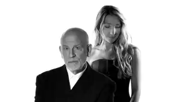 A man in a suit, behind him stands a woman with long hair.