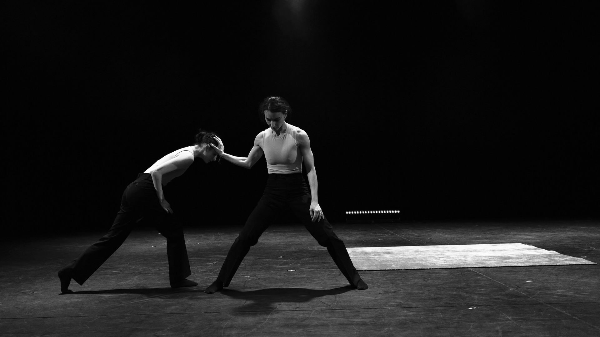 A male dancer stands with his legs wide apart, holding a hand towards a female dancer who presses her head against his hand.