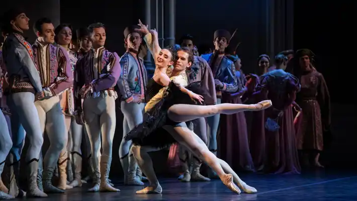 Dansare står på en scen, i förgrunden en kvinna i svarta kläder som lyfts av en manlig dansare.