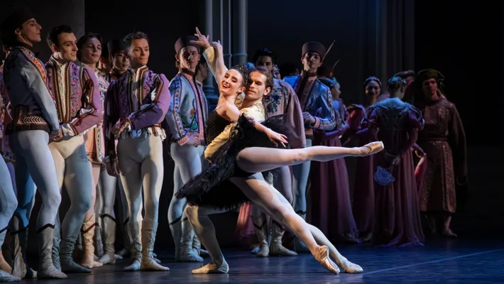 Dancers stand on a stage, in the foreground a woman in black clothes being lifted by a male dancer.