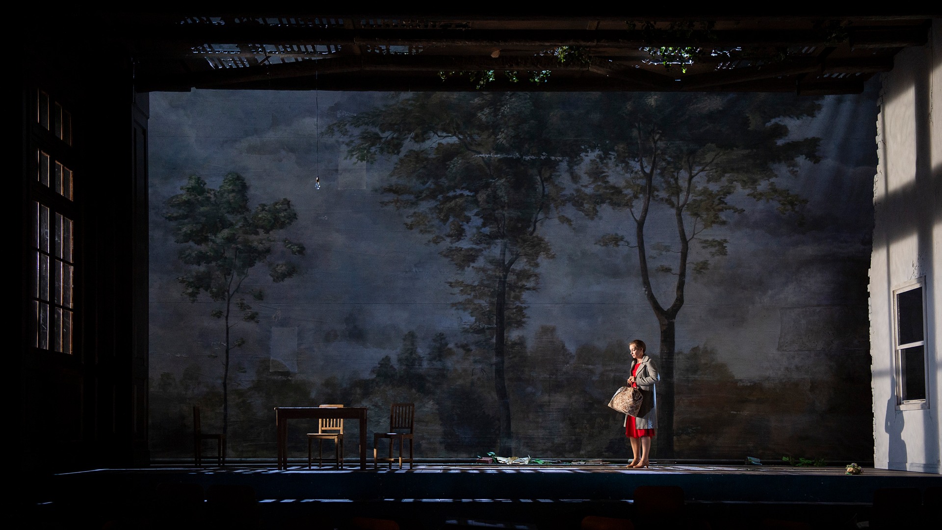 On a large stage with a landscape in the background, a lone woman in a red dress stands with a suitcase.