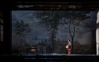 På en stor scen med ett landskap i fonden står en ensam kvinna i röd klänning med en resväska.