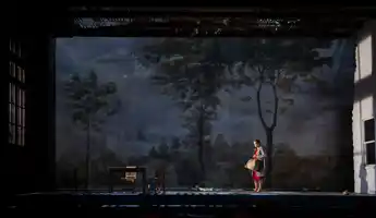 På en stor scen med ett landskap i fonden står en ensam kvinna i röd klänning med en resväska.