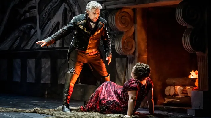 A woman lies on the floor in front of a fire, a man stands leaning over her.