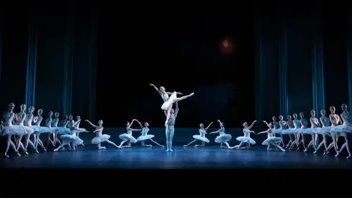 A formation of dancers in white tutus on a stage: in the center, a male dancer holds up a ballerina.