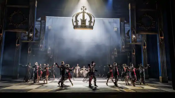 En scenbild med dekor som lyfter från golvet, en krona svävar över dansarna som rör sig i en ring.