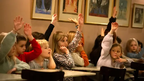 A group of children sitting in a classroom with their hands up.
