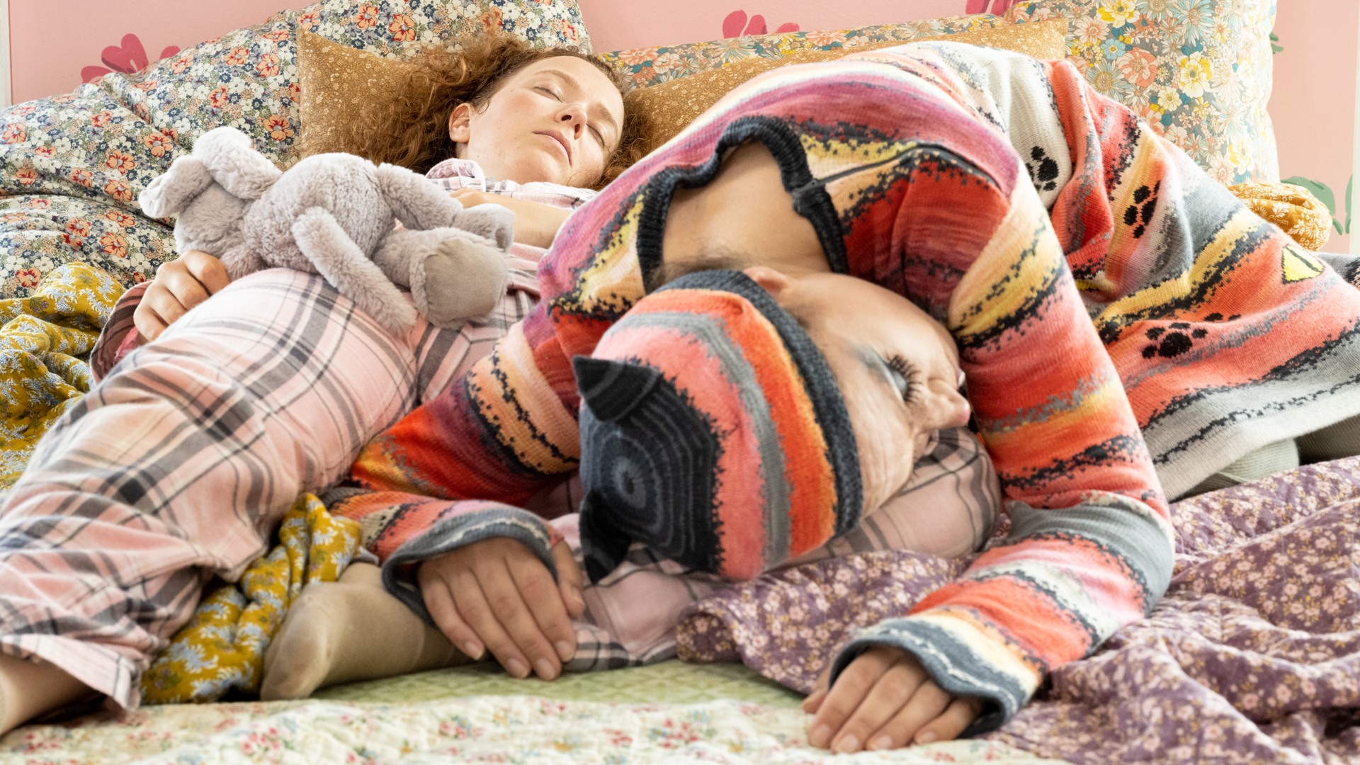 Two people lie down and sleep, blankets, pillows and stuffed animals are with them in bed.