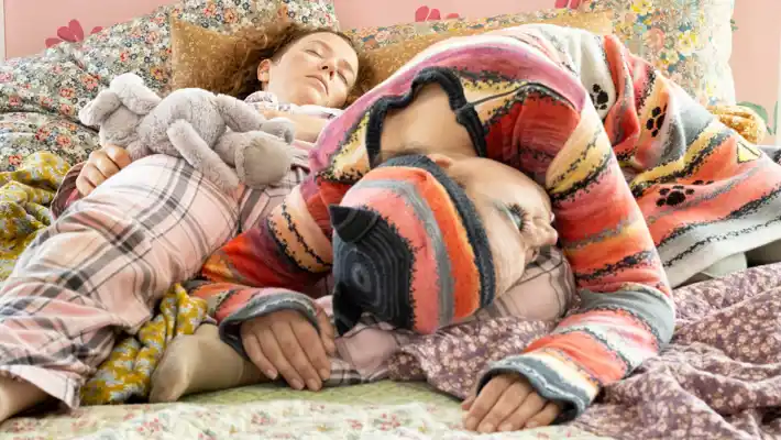 Two people lie down and sleep, blankets, pillows and stuffed animals are with them in bed.