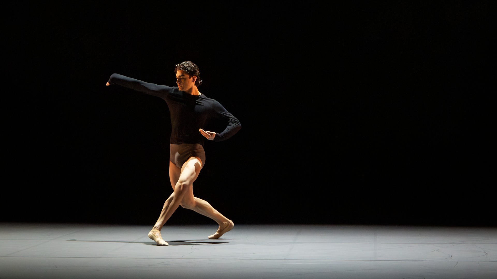 A lone dancer moves across a monochrome stage space.