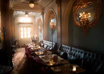 A dark room with chairs and tables, the sun shining through a window in the background.