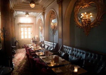 Ett mörkt rum med stolar och bord, solen lyser genom ett fönster i bakgrunden.