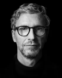 En person iklädd en mörk tröja står mot en svart bakgrund. Ansiktet är delvis i mörker, vilket gör det svårt att urskilja detaljer.