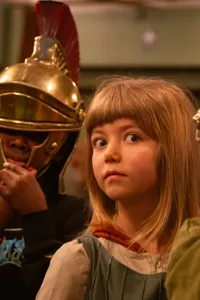 Two people in Roman helmets, their faces hidden, between them a girl with a surprised expression.