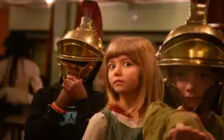 Två personer i romerska hjälmar, ansiktena dolda, mellan dem en flicka med förvånad min.