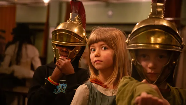 Two people in Roman helmets, their faces hidden, between them a girl with a surprised expression.