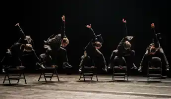 Fem dansare i kostymer hoppar på stolar.