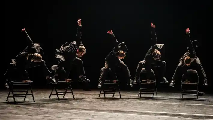 Fem dansare i kostymer hoppar på stolar.