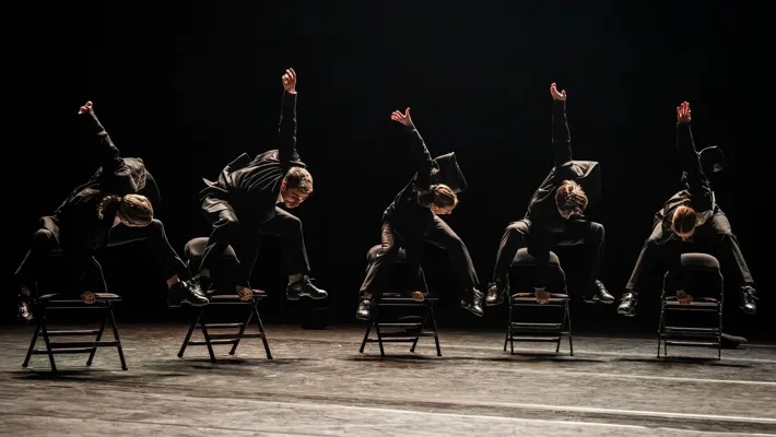 Fem dansare i kostymer hoppar på stolar.