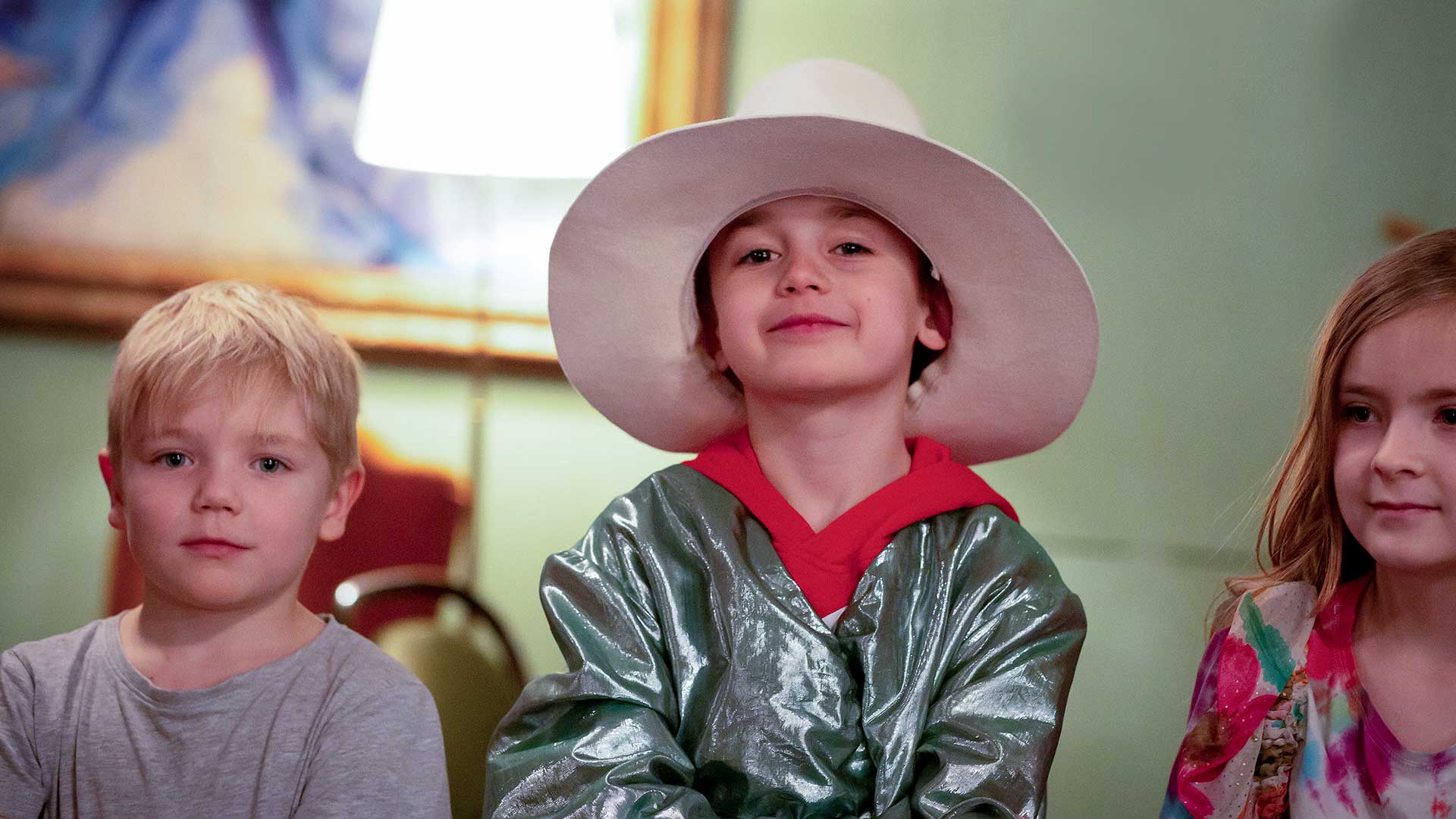 Three younger children in a row, the child in the middle has a hat and is dressed up.