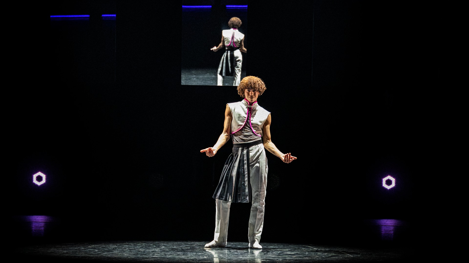 En ensam dansare står på en scen, i bakgrunden syns samma dansare på en skärm.