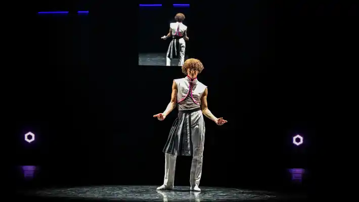 En ensam dansare står på en scen, i bakgrunden syns samma dansare på en skärm.