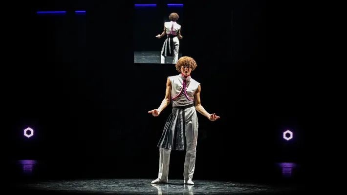 En ensam dansare står på en scen, i bakgrunden syns samma dansare på en skärm.