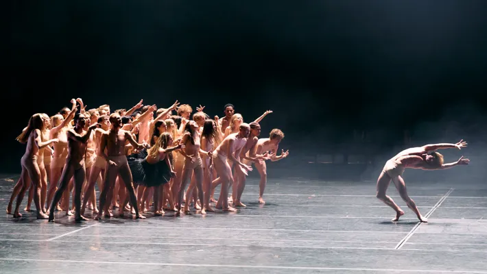 On an empty stage, a large group of dancers in skin-colored clothing stands on the left, a lone dancer on the right.