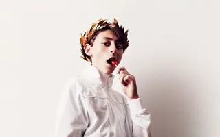 Ung person i vit skjorta med lagerkrans på huvudet och klubba i munnen mot en ljus bakgrund.