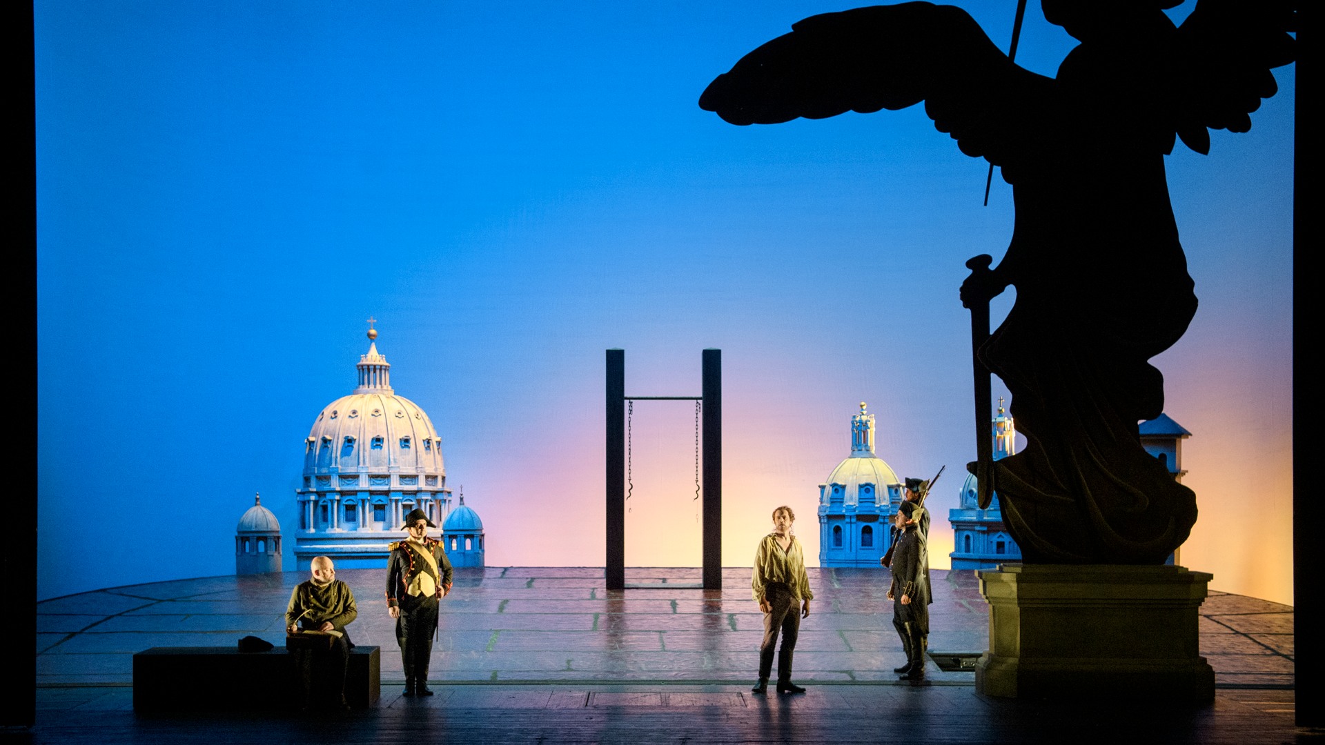 An open space with a hanging device, several soldiers and a lost man, in the background a blue sky and house facades.