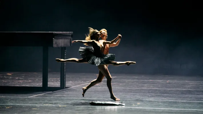 Two dancers move across an empty stage, the woman in a jump, in front of them a fish lies on the floor.