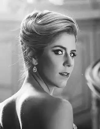 A close-up of a blonde woman, her back turned to the camera, she looks over her shoulder at us.