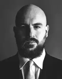 En närbild på en man, han har skägg och mustasch, är klädd i kostym och bär slips.