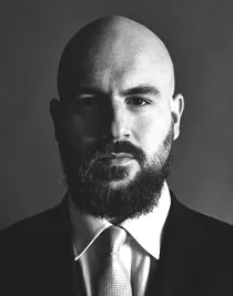 En närbild på en man, han har skägg och mustasch, är klädd i kostym och bär slips.