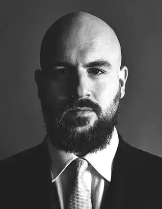 A close-up of a man, he has a beard and mustache, is dressed in a suit and wears a tie.