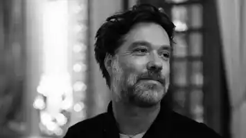 A black and white image of a man with dark hair and a grey beard in an indoor setting.