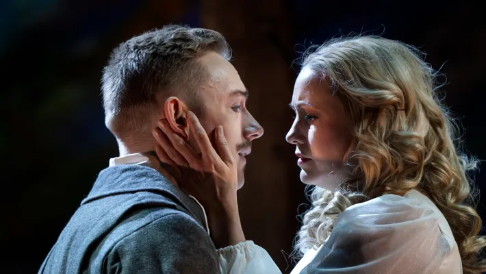 A man and a woman standing opposite each other, she holds her hand against his cheek.